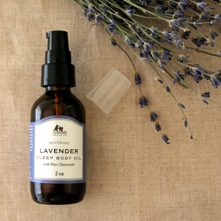 A bottle of Lavender Sleep Body Oil with a spray dispenser, accompanied by a bar of soap and lavender flowers in the background. 123 Farm is available at Luca Essentials in Los Angeles.