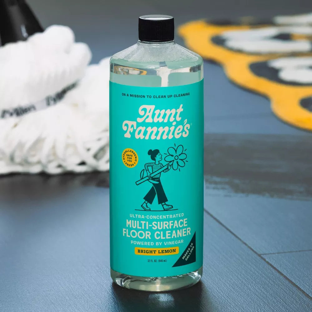 A bottle of Aunt Fannie's Multi-Surface Floor Cleaner with a blue label is on a vinyl floor with a mop in the background. Confidently mop your floors with Aunt Fannie's natural floor cleaner, available at Luca Essentials in Los Angeles.