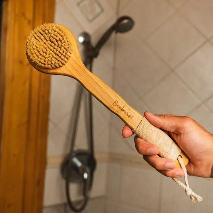 A beige bamboo dry brush with sisal bristles being held by a hand, with a shower head in the background. Bamboo Switch is available at Luca Essentials in Los Angeles.