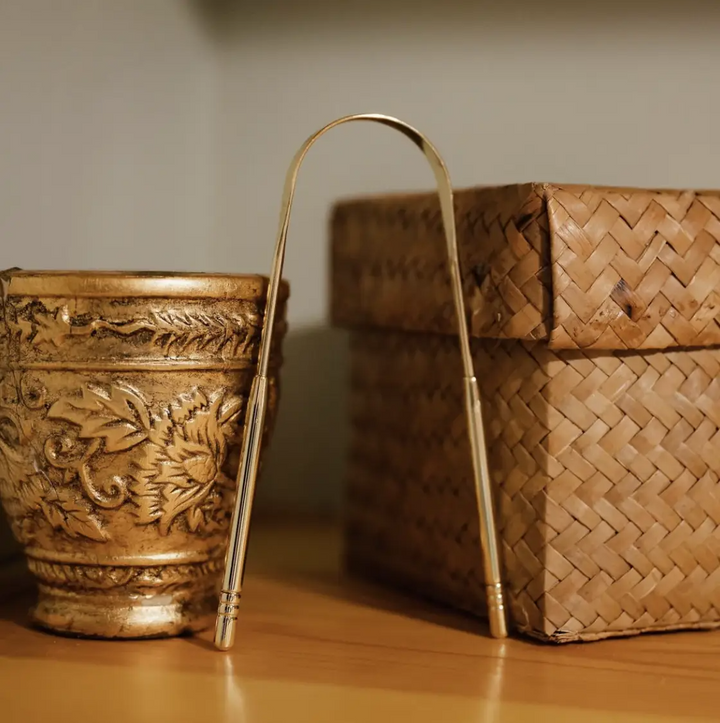 A U-shaped gold tongue scraper stands between an ornate gold cup and a woven box. Bamboo Switch is available at Luca Essentials in Los Angeles.