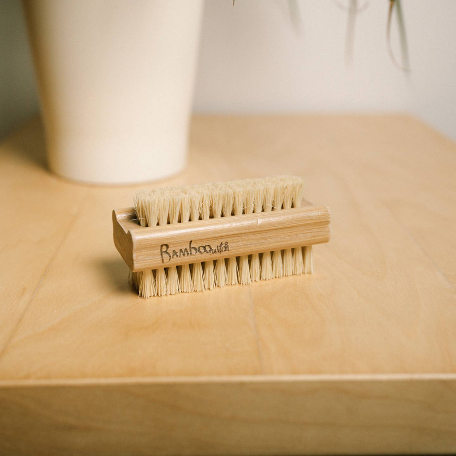 A double sided bamboo nail brush is on a wooden table with a white background. Bamboo Switch is available at Luca Essentials in Los Angeles.