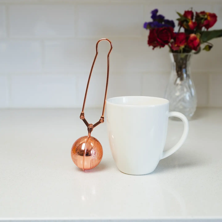 Copper tea ball infuser next to a white mug on a light surface with flowers in the background. Bamboo Switch is available at Luca Essentials in Los Angeles.