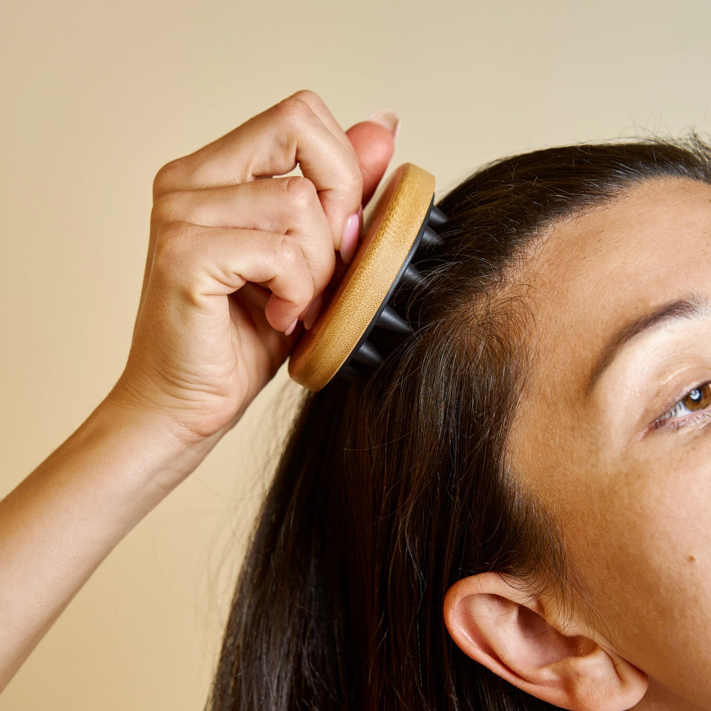 Person using a wooden hairbrush on their head against a beige background. Bamboo Switch is available at Luca Essentials in Los Angeles.