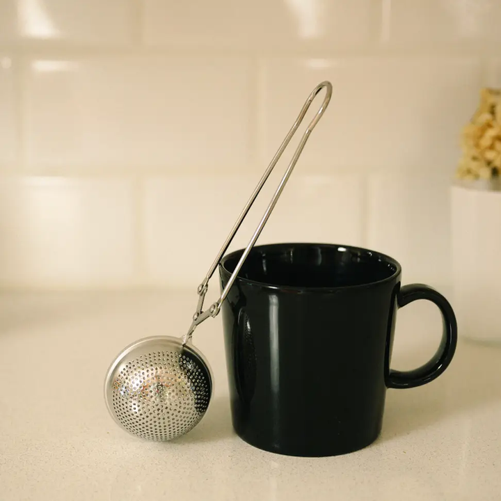 Black mug with a silver tea ball strainger on a beige surface. Bamboo Switch is available at Luca Essentials in Los Angeles.