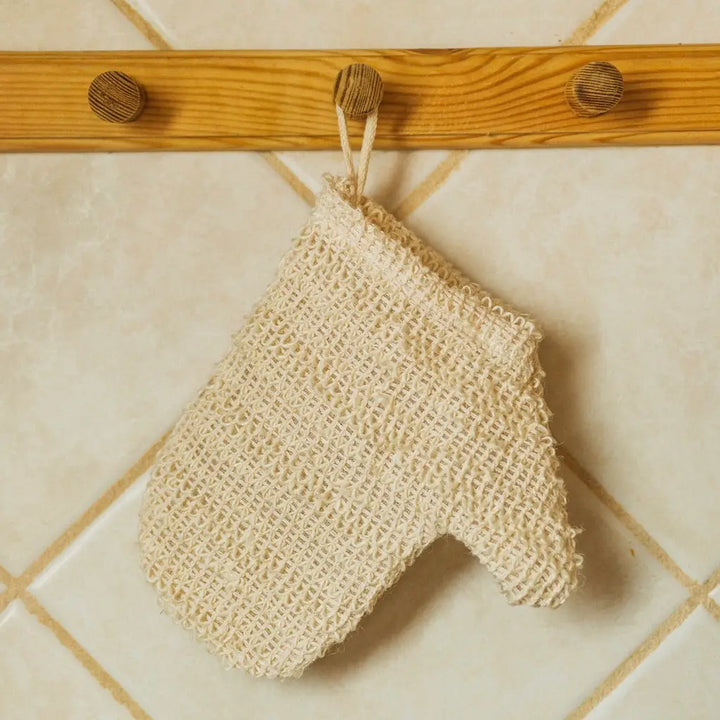 A beige sisal exfoliating shower glove hanging on a wooden hook against a tiled wall background. Bamboo Switch is available at Luca Essentials in Los Angeles.
