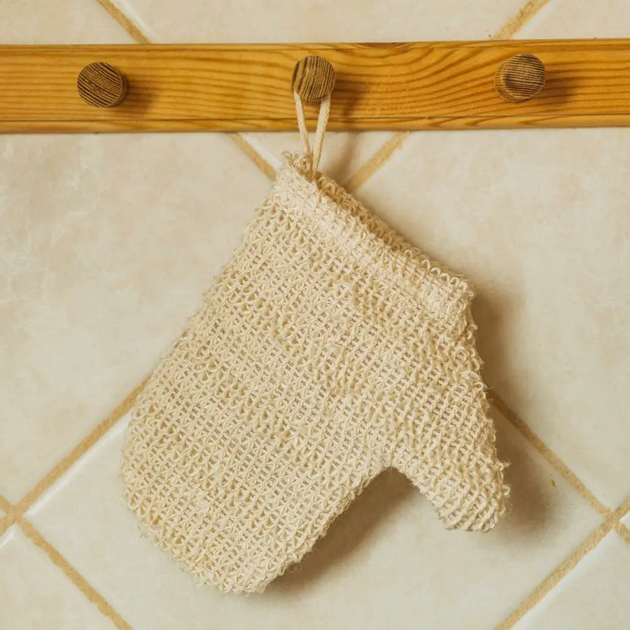 A beige sisal exfoliating shower glove hanging on a wooden hook against a tiled wall background. Bamboo Switch is available at Luca Essentials in Los Angeles.