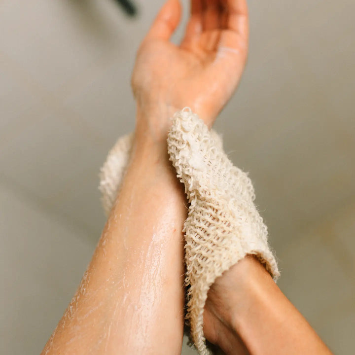 A hand cleans the other arm with a beige sisal exfoliating shower glove hanging on a wooden hook against a tiled wall background. Bamboo Switch is available at Luca Essentials in Los Angeles.