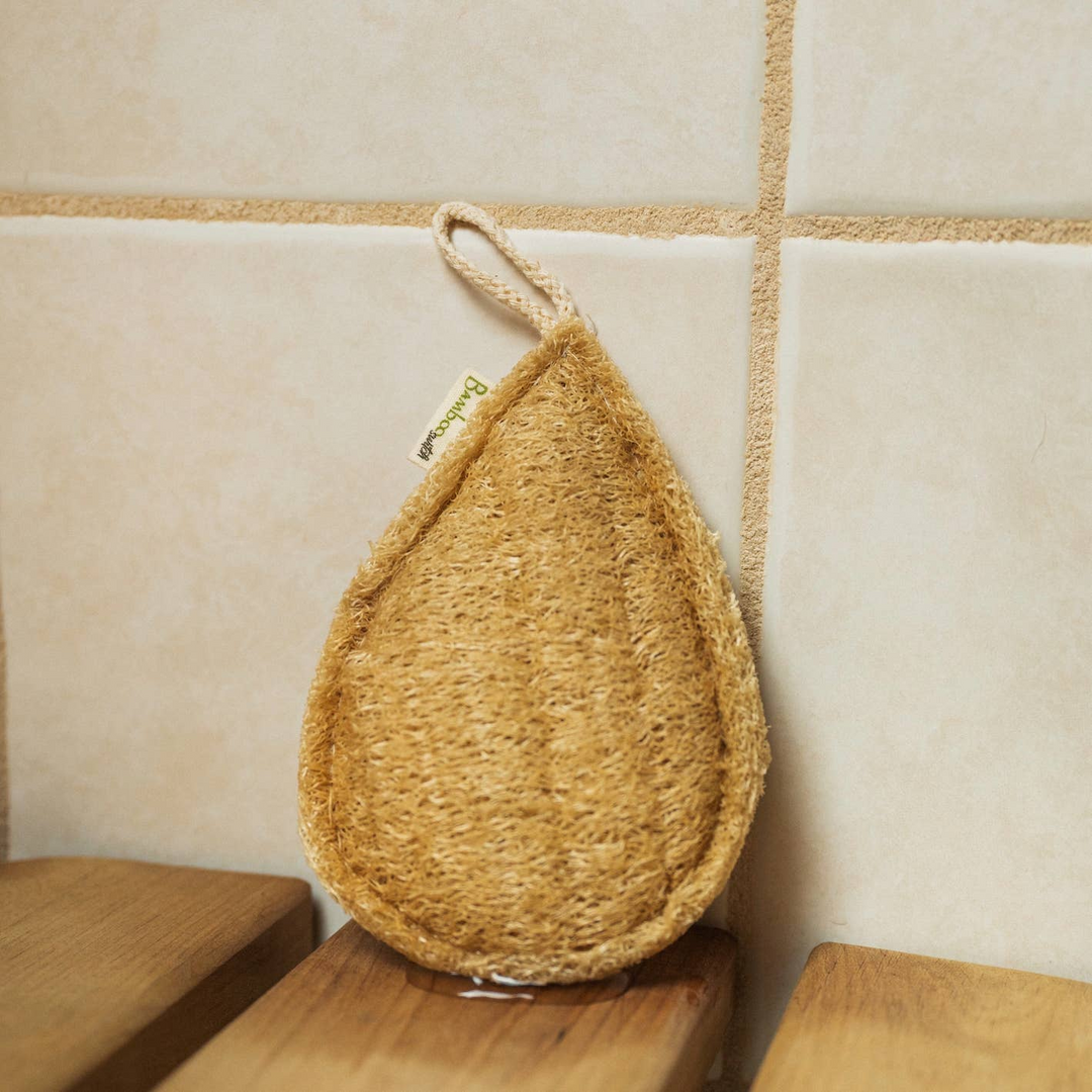 A teardrop-shaped loofah hanging on a wall hook, with a beige/natural color, against a tiled wall background. Bamboo Switch is available at Luca Essentials in Los Angeles.