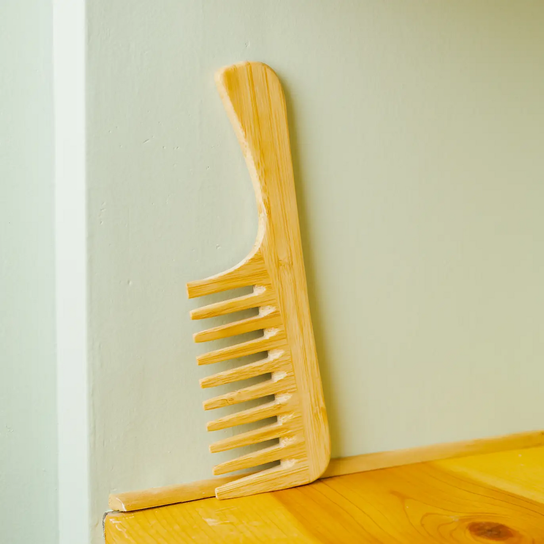 A natural light brown bamboo wide detangling comb placed against a pale green wall on a wooden surface. Bamboo Switch is available at Luca Essentials in Los Angeles.