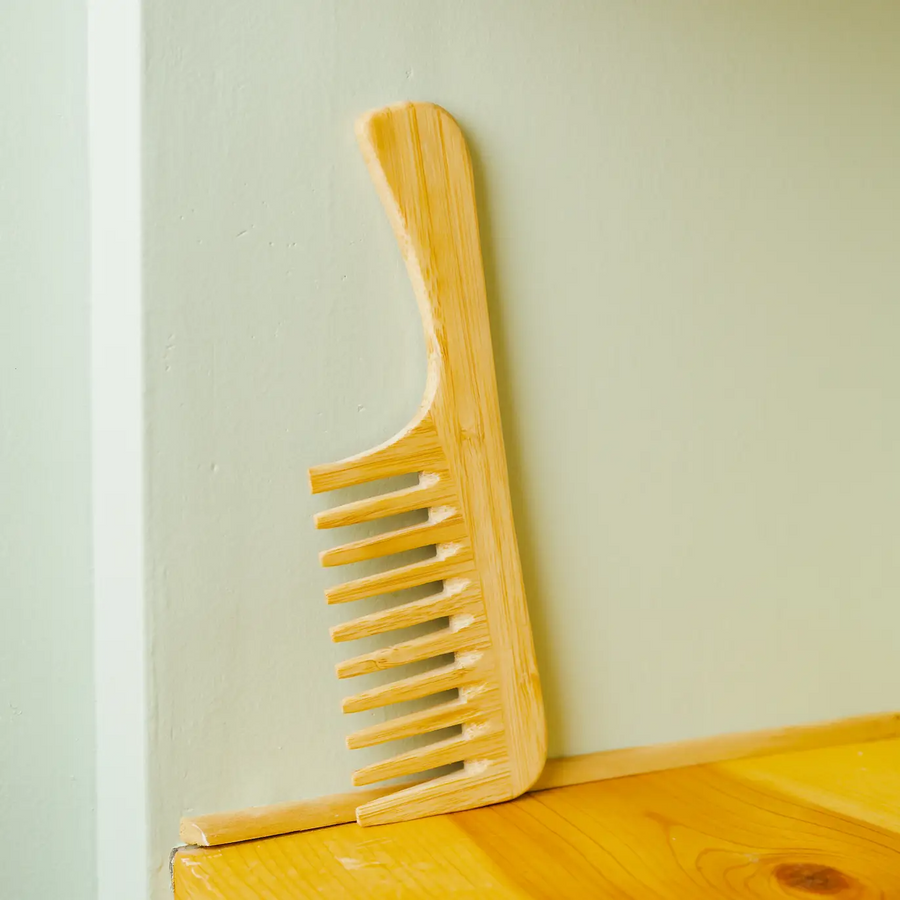 A natural light brown bamboo wide detangling comb placed against a pale green wall on a wooden surface. Bamboo Switch is available at Luca Essentials in Los Angeles.