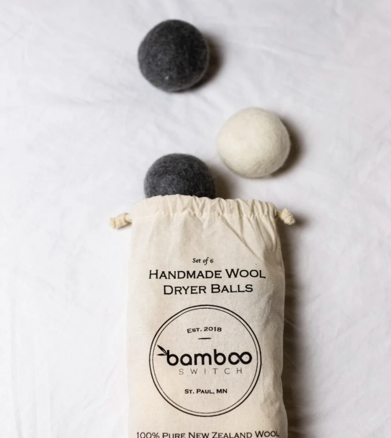 An organic cotton bag labeled 'Bamboo Switch' with a graphic of three white and three grey handmade wool dryer balls spilling out. The bag is resting on a light-colored surface with two grey balls and one white ball visible outside the bag. Bamboo Switch is available at Luca Essentials in Los Angeles.
