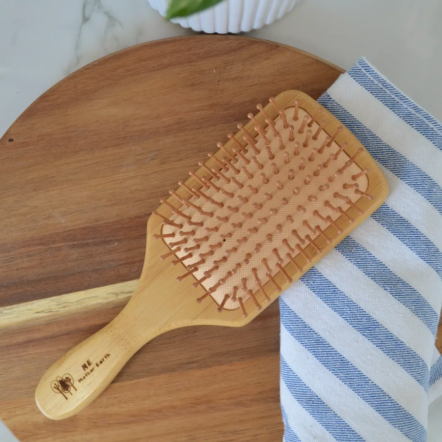 A bamboo hairbrush with a wide paddle laying on a wooden surface next to a striped towel. Bamboo Switch is available at Luca Essentials in Los Angeles.