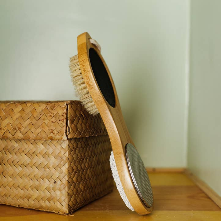 A bamboo pedicure brush with a metal file and a pumice stone attached, resting on a wicker surface. Bamboo Switch is available at Luca Essentials in Los Angeles.