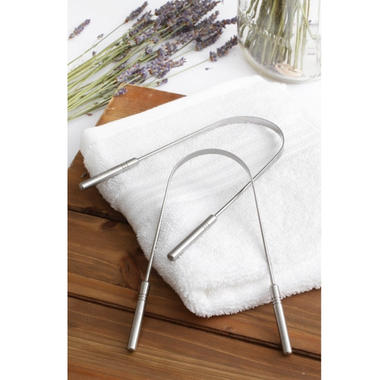Two U-shaped stainless steel tongue scrapers resting on a white towel on wooden table with dried lavender in the background. Bamboo Switch is available at Luca Essentials in Los Angeles.