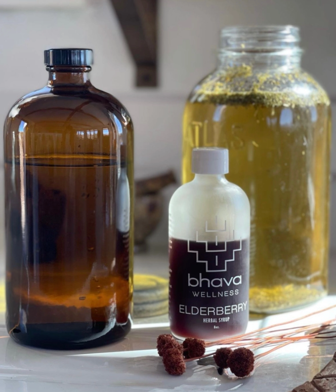 A series of glass bottles on a kitchen counter, with the foreground bottle labeled "Bhava Wellness Elderberry Herbal Syrup" with some dried flowers in the forefront. Bhava Wellness is available at Luca Essentials in Los Angeles. 