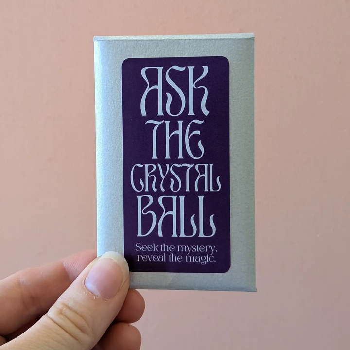 A hand holding a silver rectangular card or box with a large purple label that reads 'ASK THE CRYSTAL BALL' and smaller text below, against a soft pink background. Available at Luca Essentials in Los Angeles. 