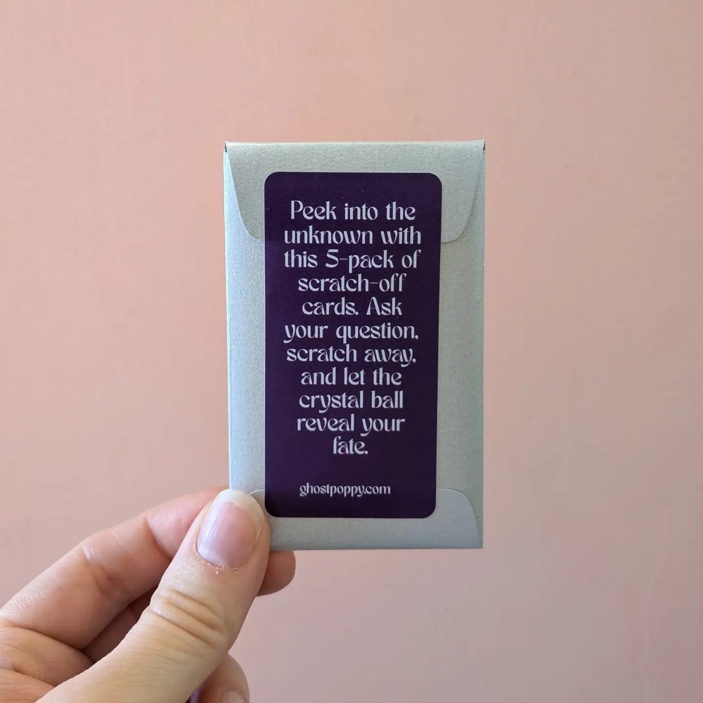 A hand holds a small gray envelope-style box with a dark purple label that reads, 'Peek into the unknown with this 5-pack of scratch-off cards. Ask your question, scratch away, and let the crystal ball reveal your fate'. Available at Luca Essentials in Los Angeles. 