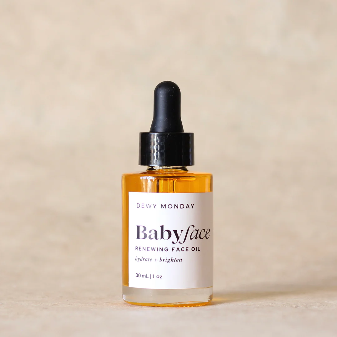 A bottle of Dewy Monday Babyface Renewing Face Oil with amber liquid, a white label, and a black dropper cap, set against a warm beige textured background. Available at Luca Essentials in Los Angeles. 