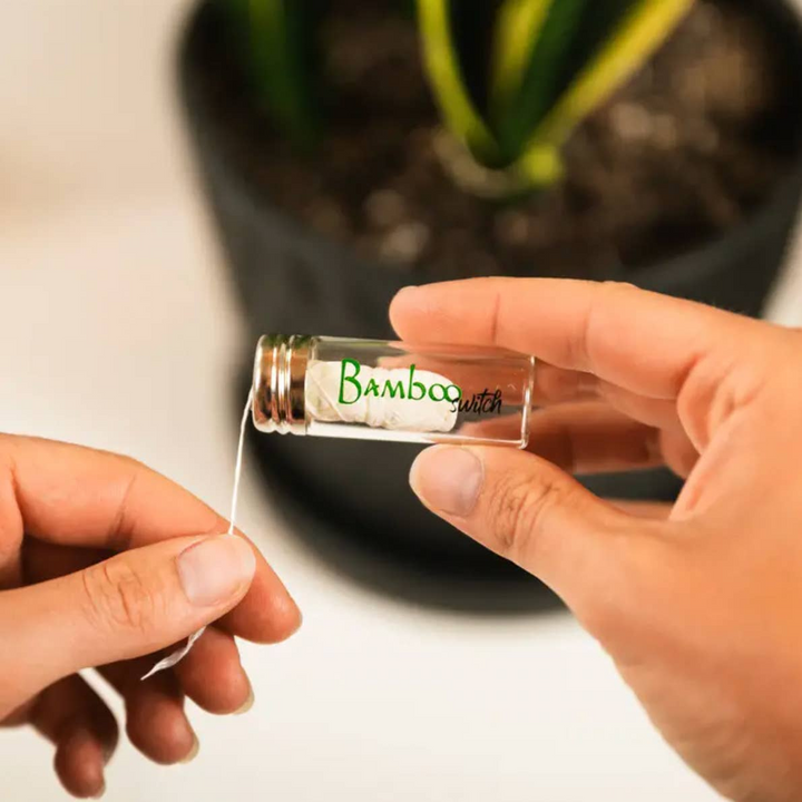 A clear glass bottle with a metal cap, labeled 'Bamboo Switch', being held by a person with visible hands and a potted plant in the background. Bamboo Switch is available at Luca Essentials in Los Angeles.