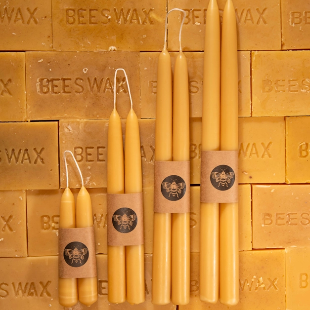 Four natural beeswax taper candles in different sizes, wrapped in brown paper bands with a circular bee logo, arranged against a background of stacked beeswax bars. From left to right the sizes are: 6", 9", 12" and 15". Available at Luca Essentials in Los Angeles.