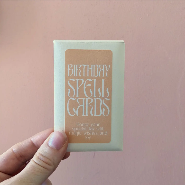 Hand holding a pack of 'Birthday Spell Cards' against a pink background. Available at Luca Essentials in Los Angeles.