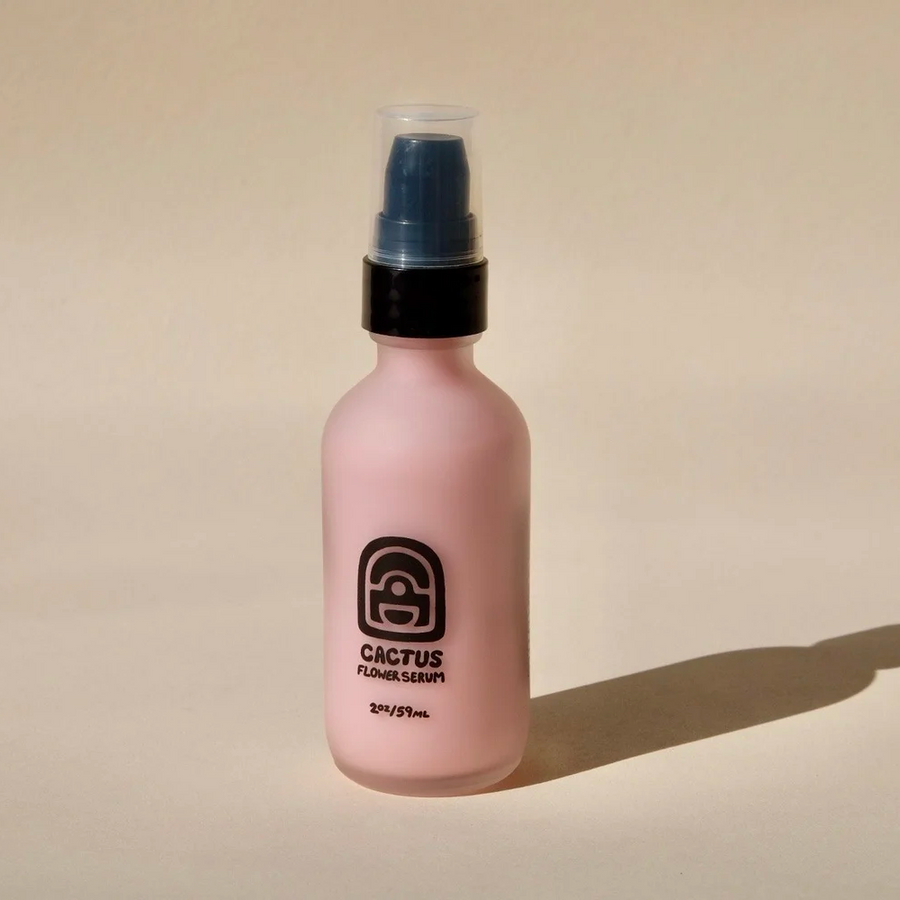 A frosted light pink bottle with a black collar and black head, labeled 'Cactus Flower Serum', with a black circular illustration, set against a warm beige background with a soft shadow. Available at Luca Essentials in Los Angeles.