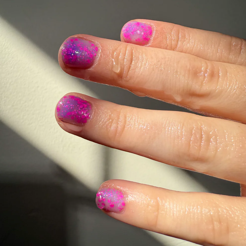 A close-up of a hand showing fingernails painted in a glittery pink and purple gradient design featuring small pink polka dots. Available at Luca Essentials in Los Angeles. 