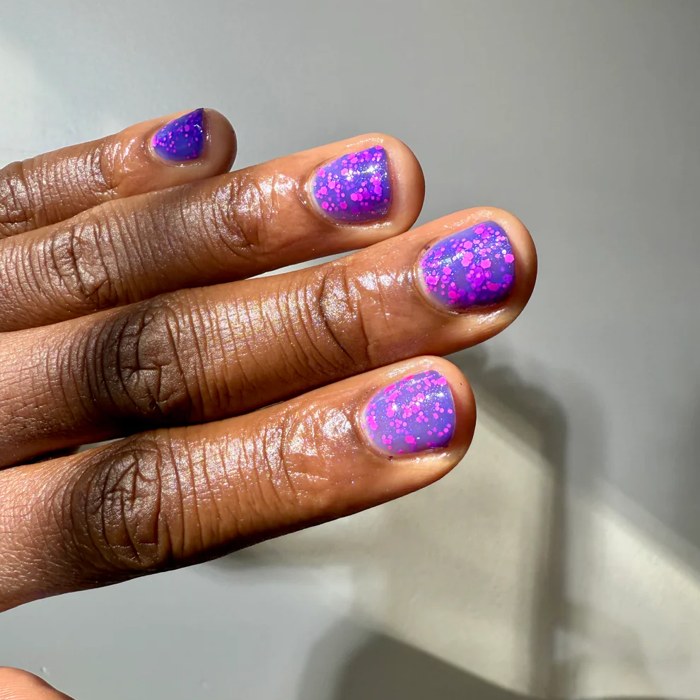 A close-up of a hand with manicured nails painted in a glittery purple and pink ombre pattern, set against a plain light gray background. Available at Luca Essentials in Los Angeles. 