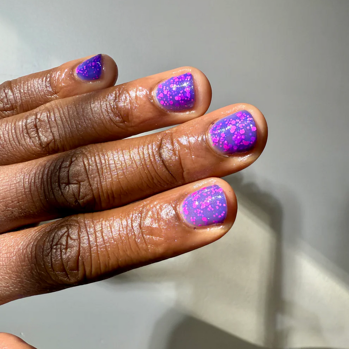 A close-up of a hand with manicured nails painted in a glittery purple and pink ombre pattern, set against a plain light gray background. Available at Luca Essentials in Los Angeles. 
