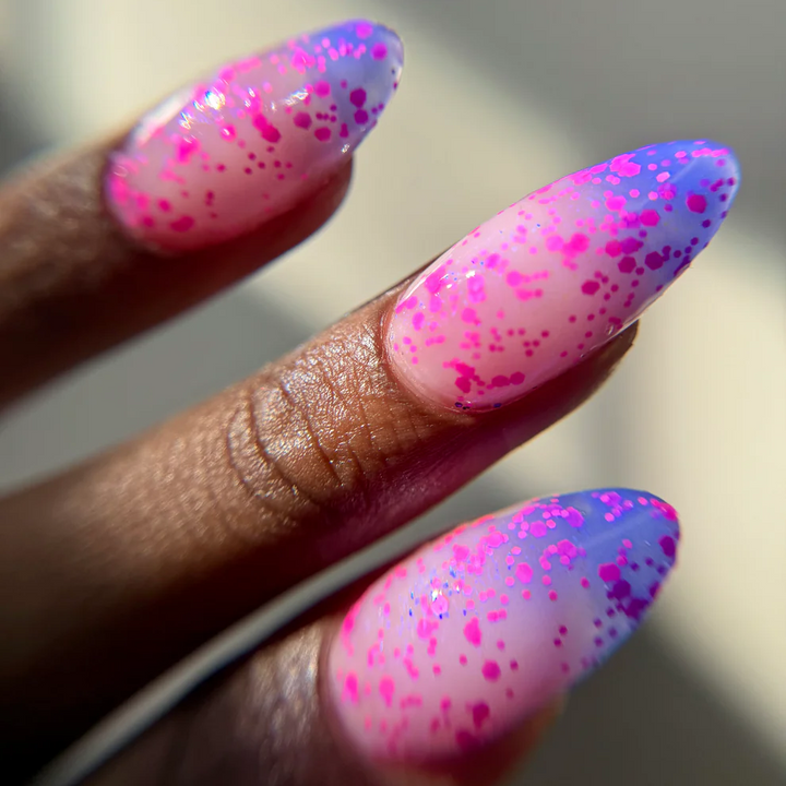 Close-up of a hand with long almond-shaped nails painted in a pink-to-blue gradient with bright pink glitter speckles. Available at Luca Essentials in Los Angeles. 