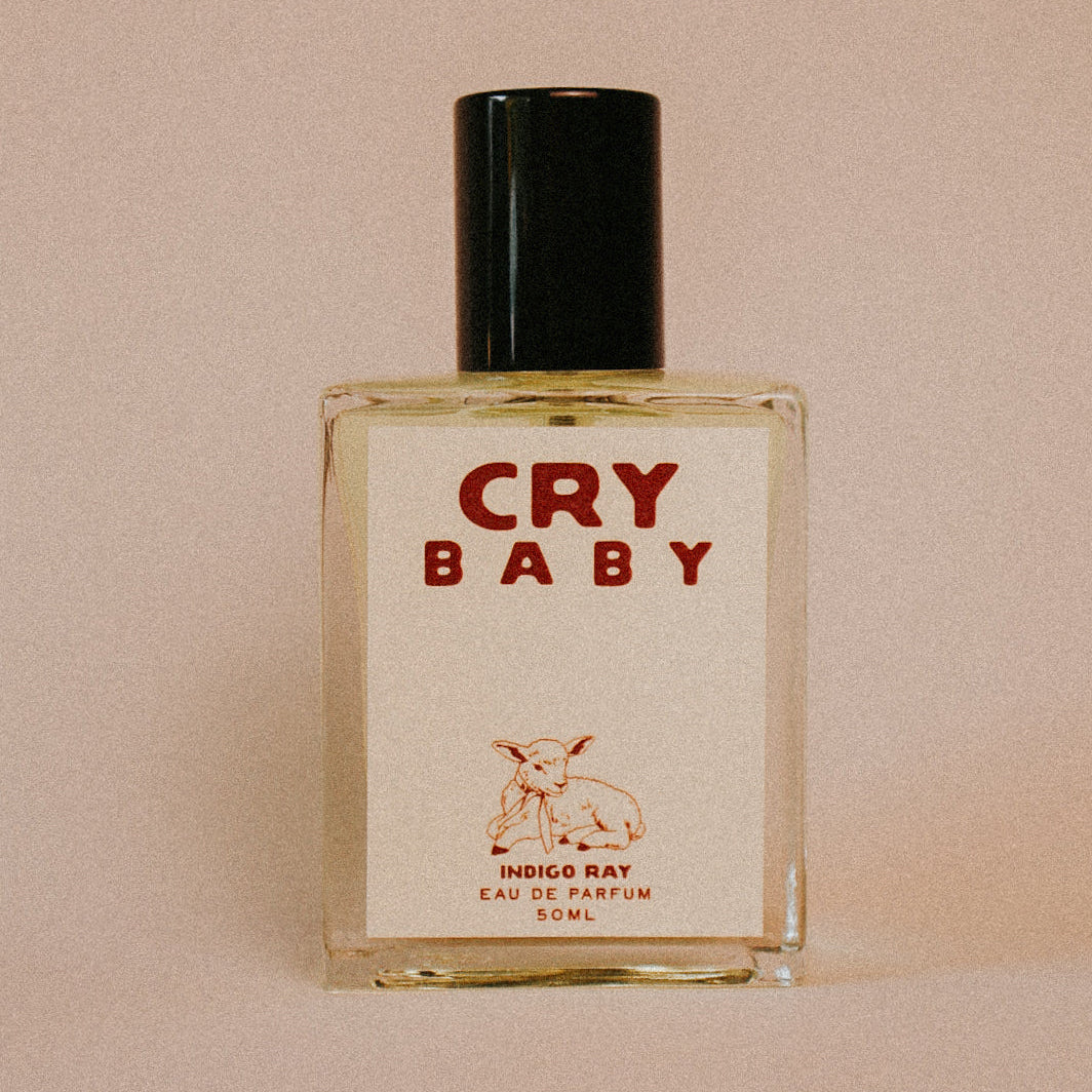 A 50ml bottle of 'Cry Baby' Eau de Parfum by Indigo Ray, featuring a square clear glass bottle with a cream-colored label and a black cap, set against a solid beige background. Available at Luca Essentials in Los Angeles. 