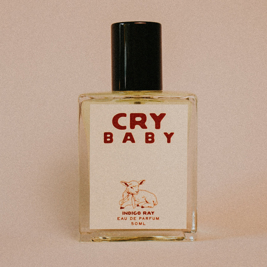 A 50ml bottle of 'Cry Baby' Eau de Parfum by Indigo Ray, featuring a square clear glass bottle with a cream-colored label and a black cap, set against a solid beige background. Available at Luca Essentials in Los Angeles. 