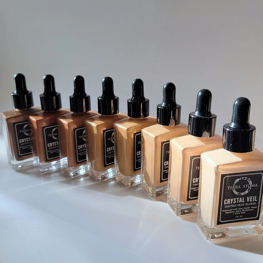 Row of 8 Terra Stoma tinted skin elixir bottles with different shades with dropper caps on a white surface. Available at Luca Essentials in Los Angeles. 