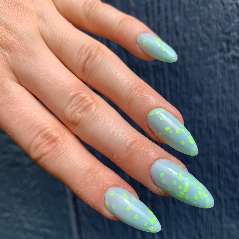 A close-up of a hand with long, almond-shaped nails painted in a pale mint green with scattered neon yellow-green polka dots, set against a dark textured background. Available at Luca Essentials in Los Angeles. 