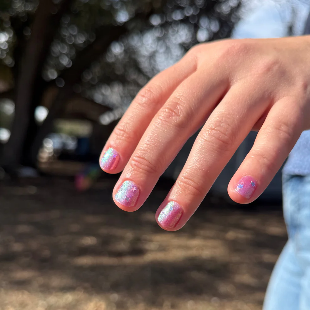 Close-up of a hand with pink glittery nail polish against a blurred natural background

Sheer pink with iridescent shimmer base thats shifts from green, teal, pale gold layered with scattered sparkly blue micro flakes and holo stars. 

Available at Luca Essentials in Los Angeles. 