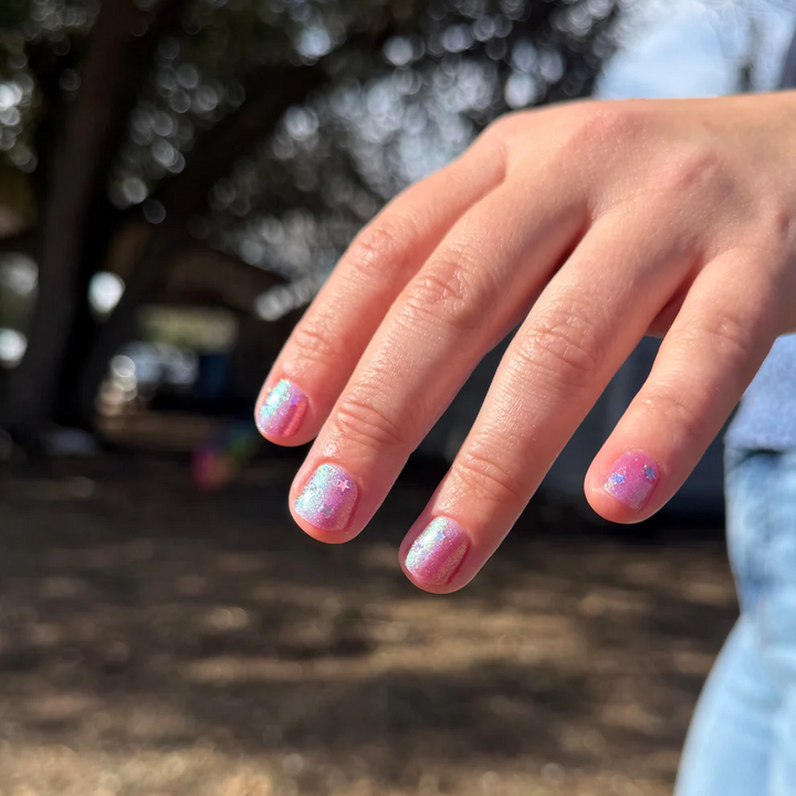 Close-up of a hand with pink glittery nail polish against a blurred natural background

Sheer pink with iridescent shimmer base thats shifts from green, teal, pale gold layered with scattered sparkly blue micro flakes and holo stars. 

Available at Luca Essentials in Los Angeles. 