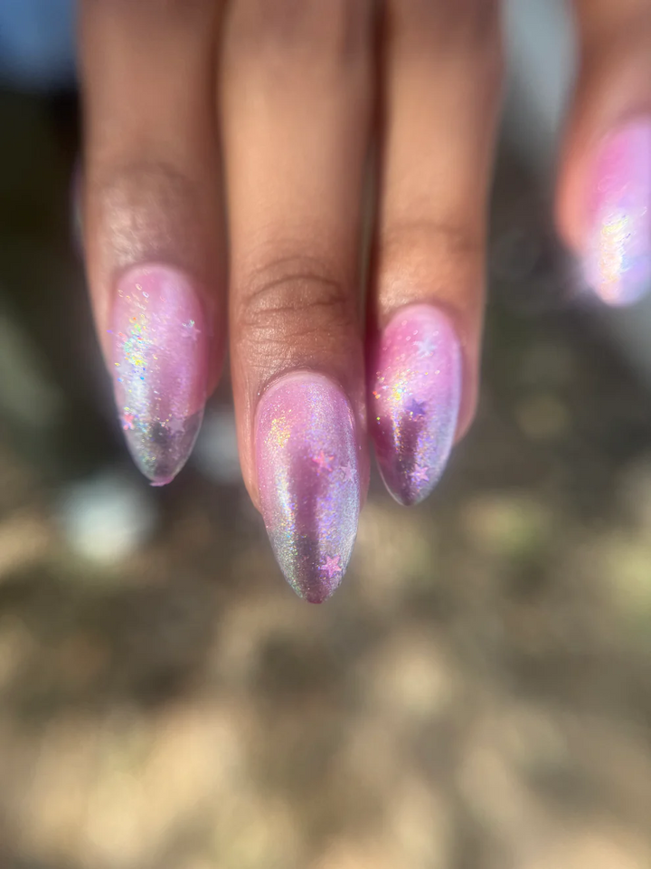 Close-up of a hand with pink and purple glittery nail polish on a blurred natural background

Sheer pink with iridescent shimmer base thats shifts from green, teal, pale gold layered with scattered sparkly blue micro flakes and holo stars. 

Available at Luca Essentials in Los Angeles. 