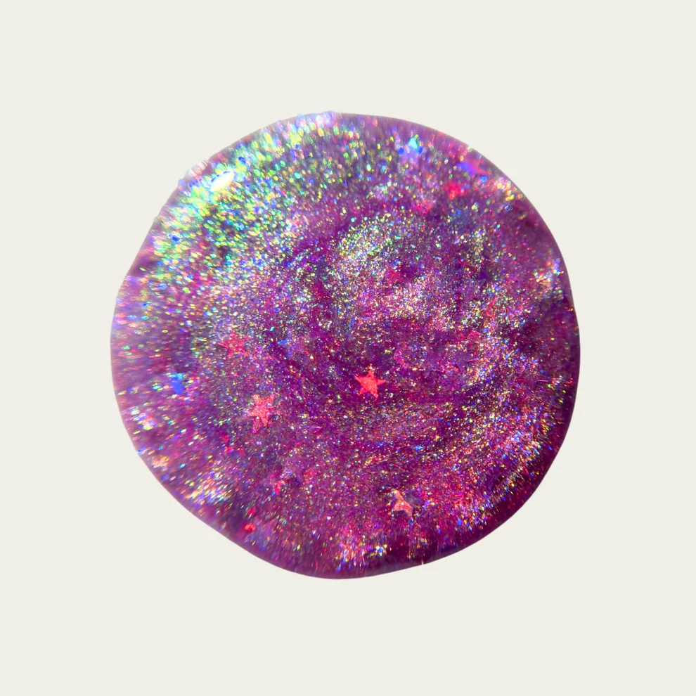 Round, glittery swatch with a pink and purple color scheme on a light gray background

Sheer pink with iridescent shimmer base thats shifts from green, teal, pale gold layered with scattered sparkly blue micro flakes and holo stars. 

Available at Luca Essentials in Los Angeles. 