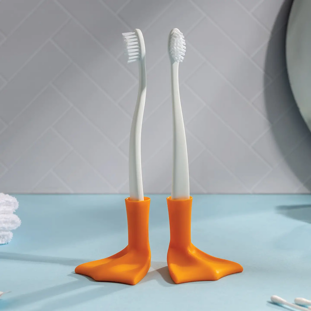Two toothbrushes with orange holders shaped like duck feet on a light blue surface. Available at Luca Essentials in Los Angeles. 