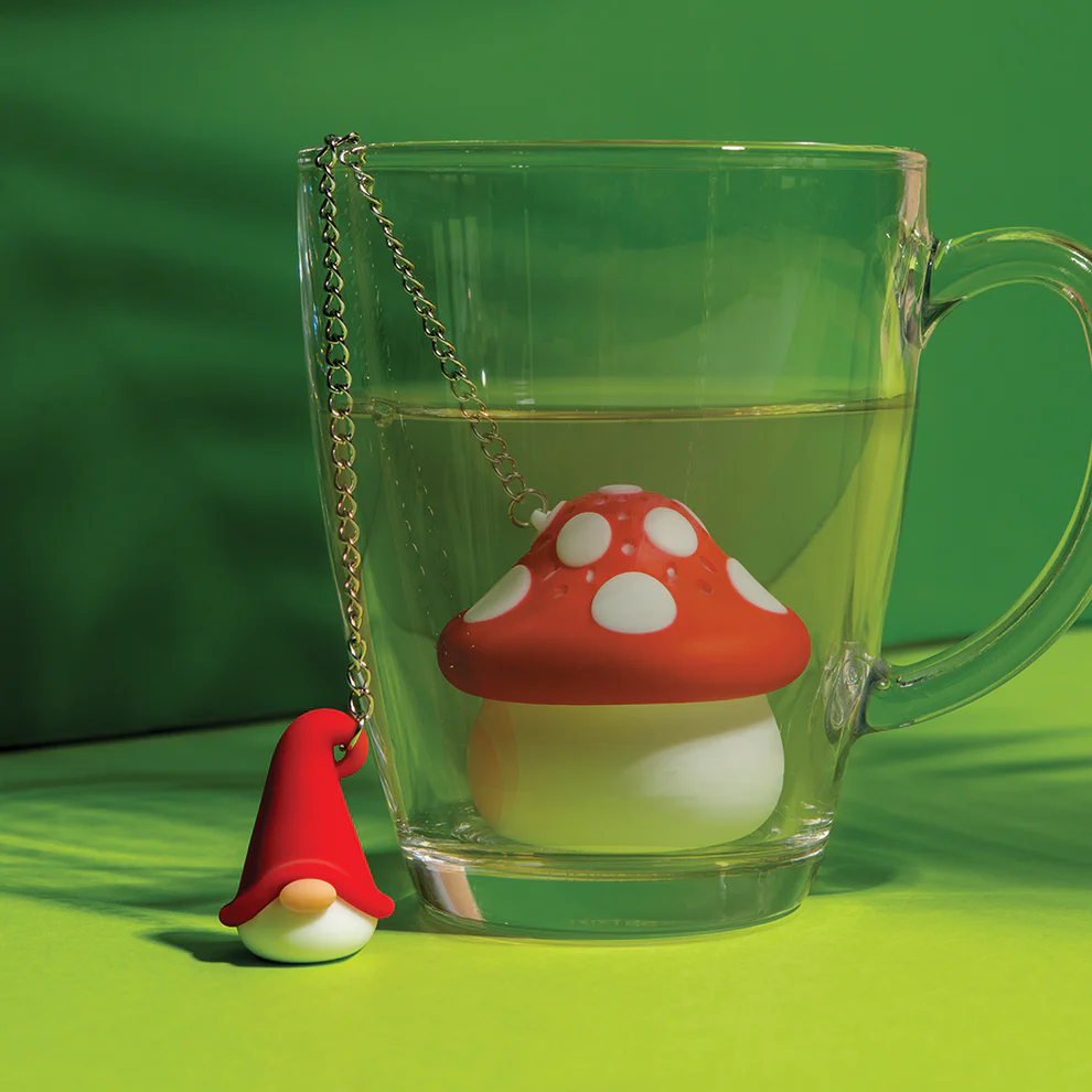 Clear mug with a red and white mushroom-shaped tea infuser  with a chain connected to a small gnome figure on a green background. Available at Luca Essentials in Los Angeles. 