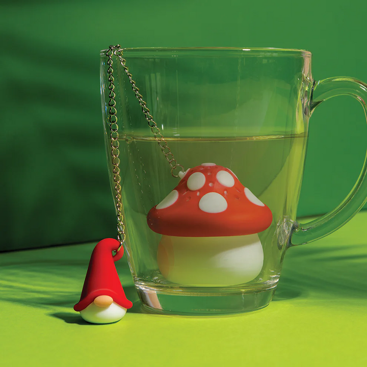 Clear mug with a red and white mushroom-shaped tea infuser  with a chain connected to a small gnome figure on a green background. Available at Luca Essentials in Los Angeles. 