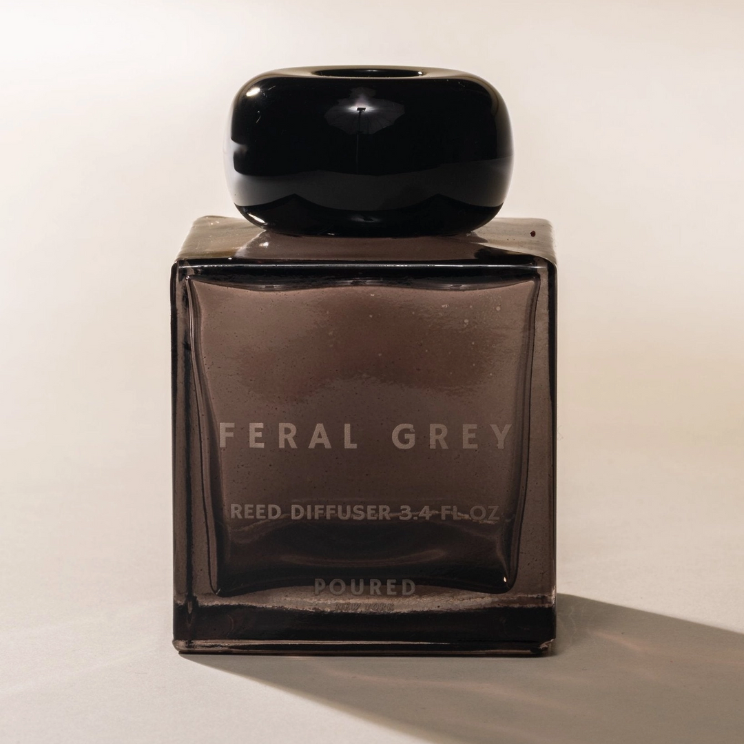 A dark grey glass cube bottle with a round black top with the title "Feral Grey" on the front on a beige background. Available at Luca Essentials in Los Angeles. 