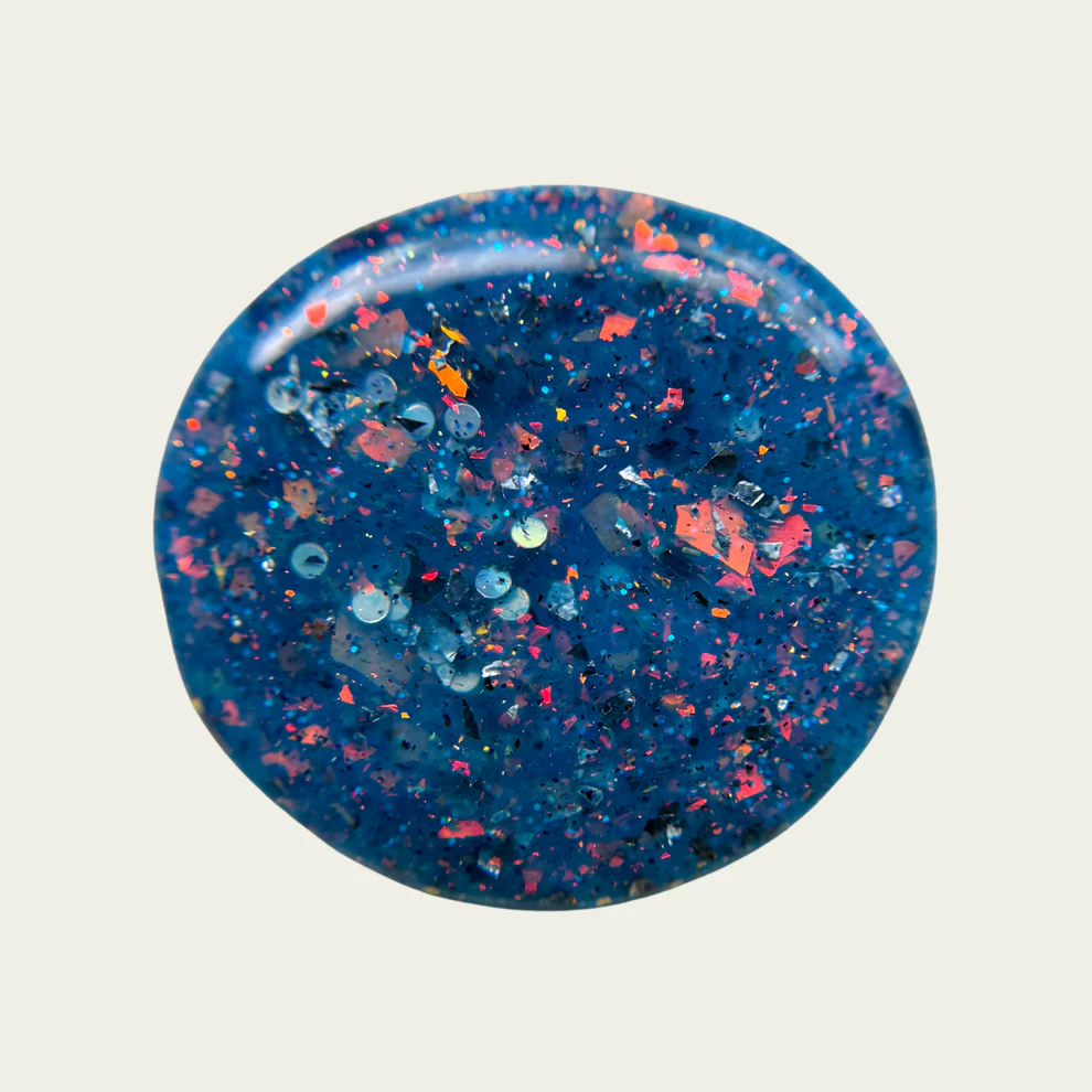 Blue speckled glitter nail polish swatch on a gray background. Available at Luca Essentials in Los Angeles. 