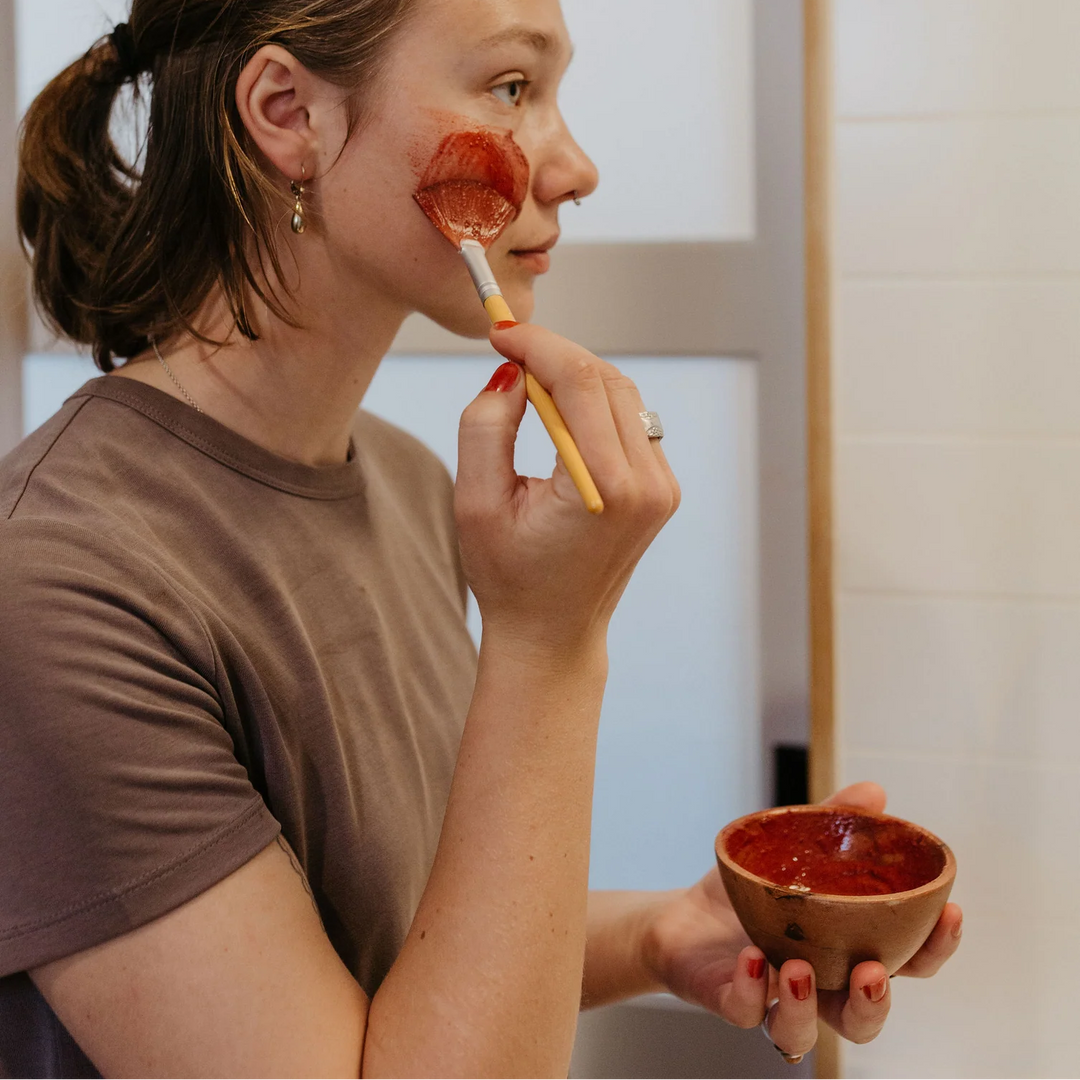 Person applying red facial mask with brush and bowl in front of mirror. Available at Luca Essentials in Los Angeles. 