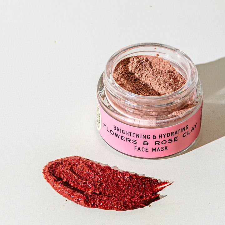 An open clear jar of clay face mask powder with a pink label reading 'Brightening & Hydrating Flowers & Rose Clay Face Mask,' placed on a white surface beside a smear of the deep rose-red powder. Available at Luca Essentials in Los Angeles. 