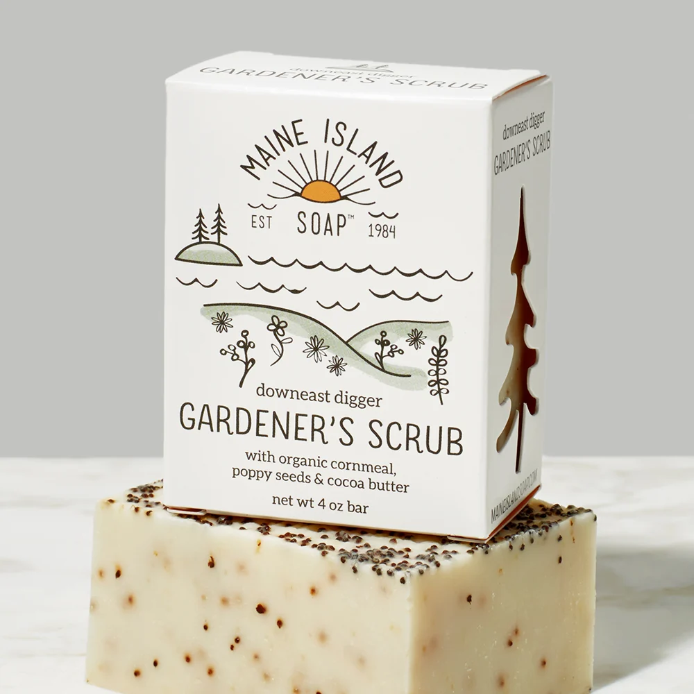 Branded soap bar in box with the logo 'Maine Island Soap' and an illustration of a mountain, trees, water and flowers. The scent 'Gardener's Scrub' is located underneath the illustration. Box of bar soap is sitting on top of a beige and brown flecked bar soap without packaging. Available at Luca Essentials in Los Angeles. 