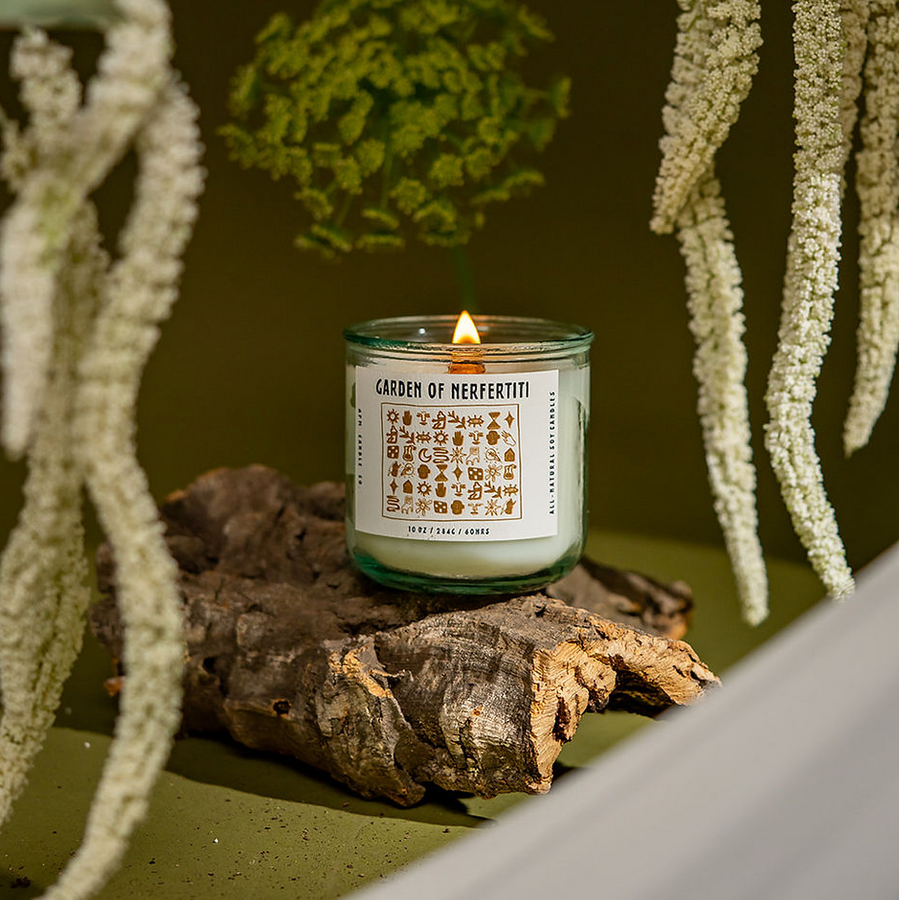 A lit candle with a white label reading "Garden of Nerfertiti" with symbols on a glass jar. The candle is lit and sitting on a piece of wood, with hanging flowers surrounding it and a green background.