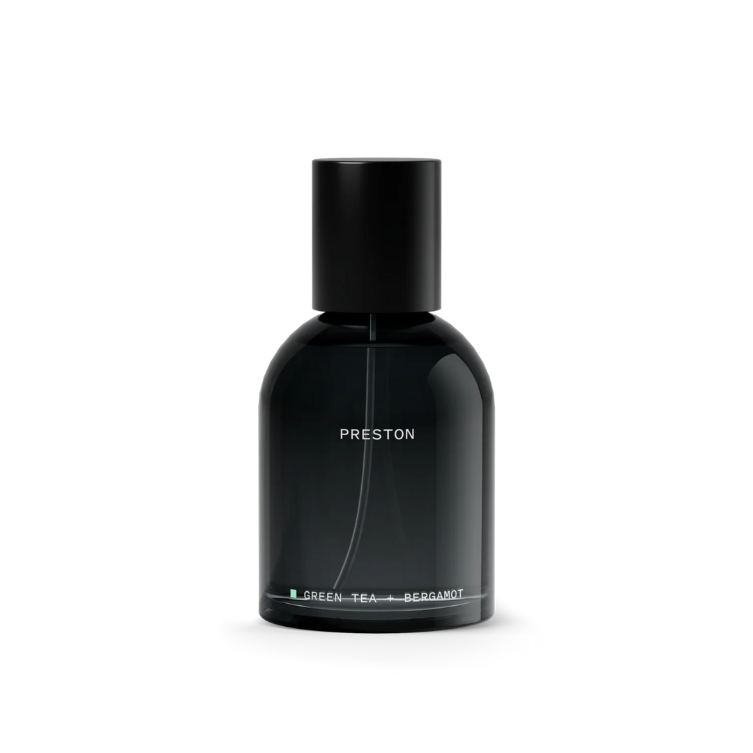 A sleek, matte black glass perfume bottle with a matching black cap, featuring white text that reads 'PRESTON' and 'GREEN-TEA + BERGAMOT' on a clean white background. Available at Luca Essentials in Los Angeles. 