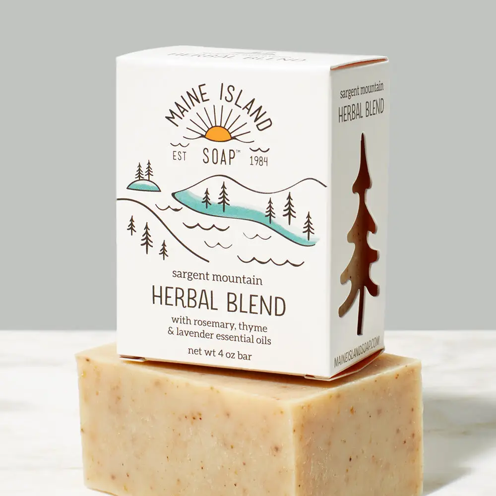 Branded soap bar in box with the logo 'Maine Island Soap' and an illustration of a mountain, trees and water. The scent 'Herbal Blend' is located underneath the illustration. Box of bar soap is sitting on top of a beige and brown flecked bar soap without packaging. Available at Luca Essentials in Los Angeles. 