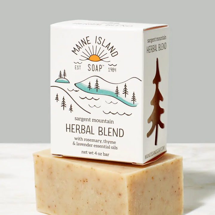 Branded soap bar in box with the logo 'Maine Island Soap' and an illustration of a mountain, trees and water. The scent 'Herbal Blend' is located underneath the illustration. Box of bar soap is sitting on top of a beige and brown flecked bar soap without packaging. Available at Luca Essentials in Los Angeles. 